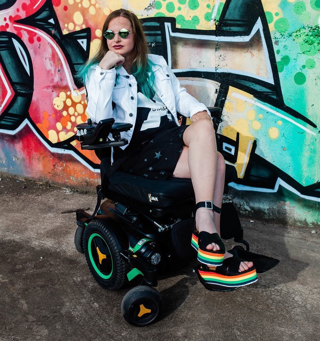 Picture of Chelsea in her wheelchair with green rims. She's wearing a youthful outfit, seated in front of a wall with colorful graffiti.