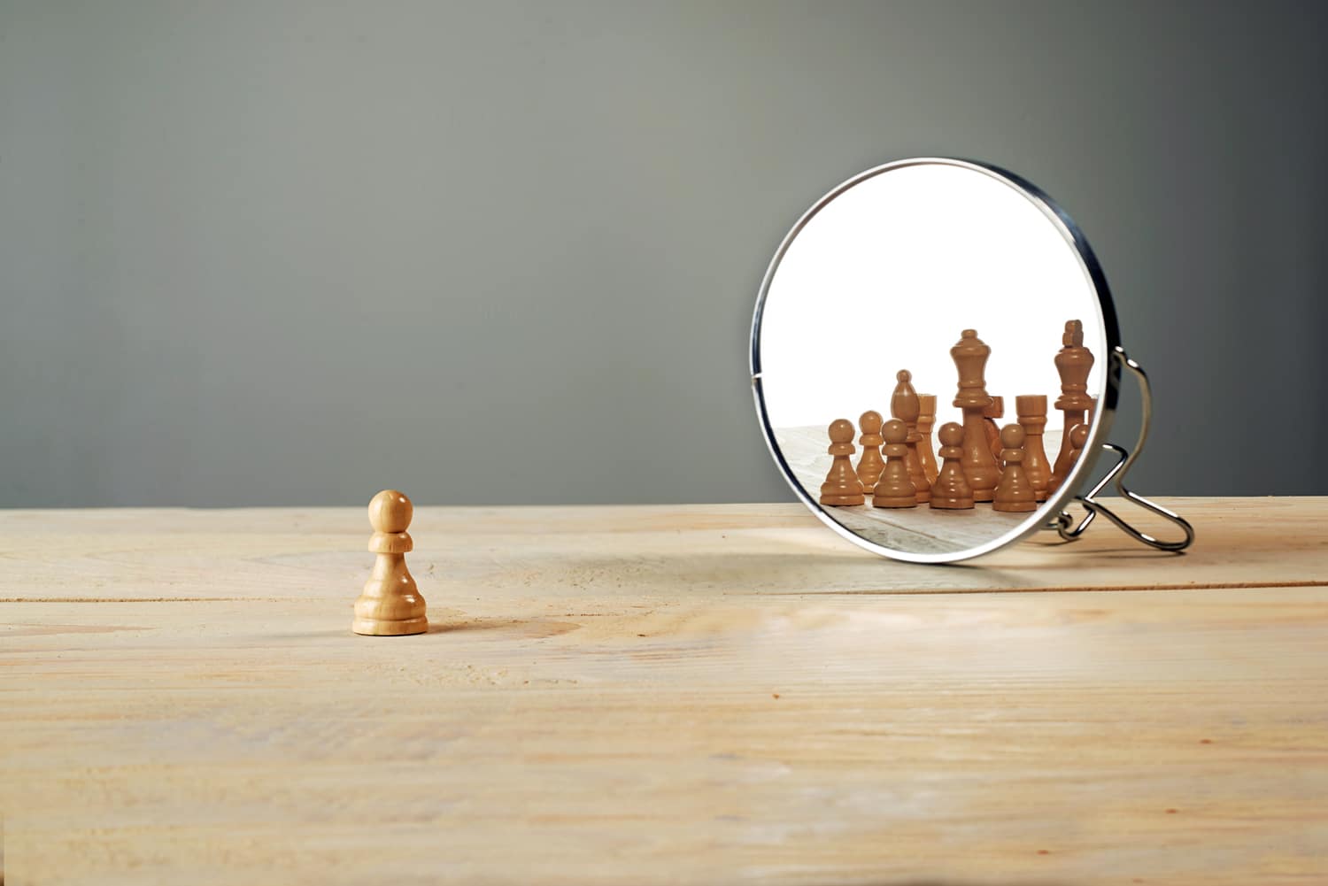 A small chess piece on a wooden table "looking" into a round mirror showing the reflection of many chess pieces, against a grey wall. 